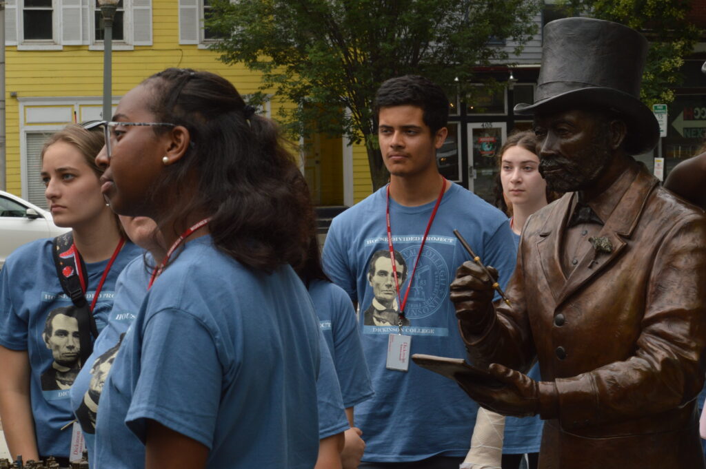 Students standing in front of a statue of a man in a nineteenth century hat and coat.