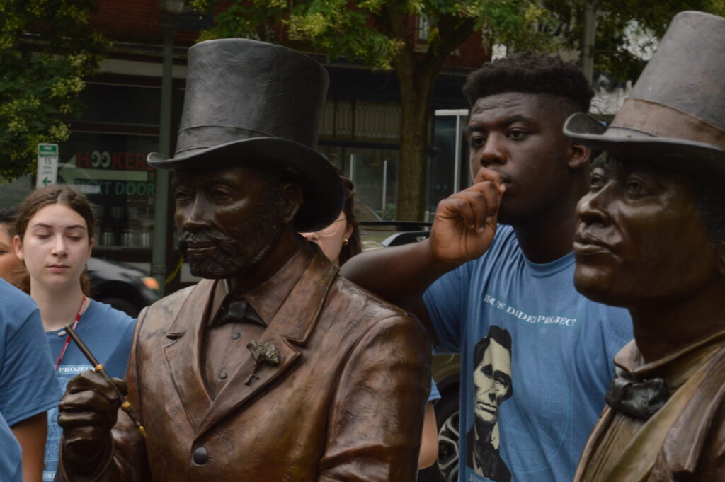 Two students standing in between two statues of people suits and hats