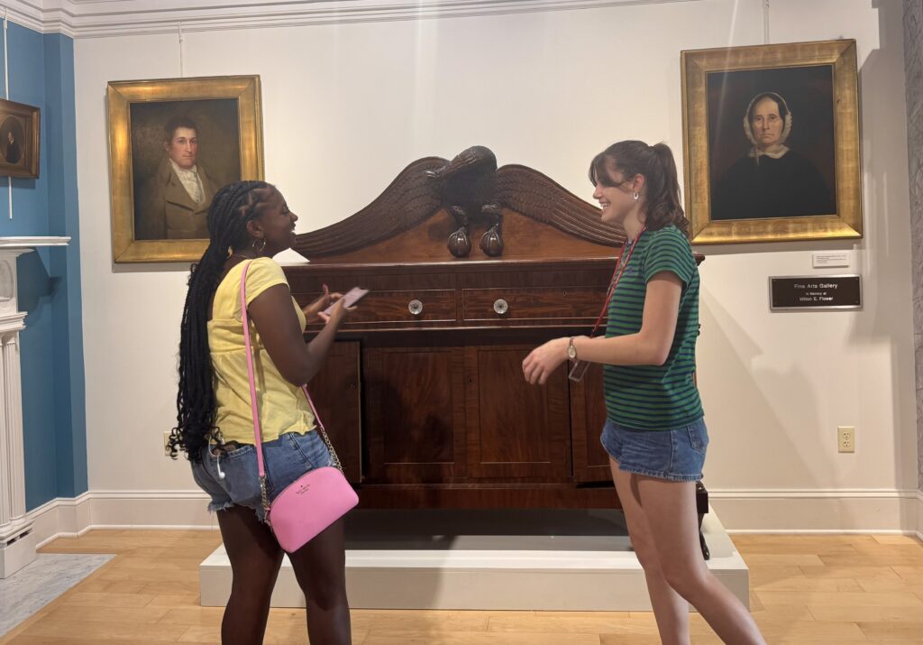 Two girls laughing in front of two paintings and a chest of drawers