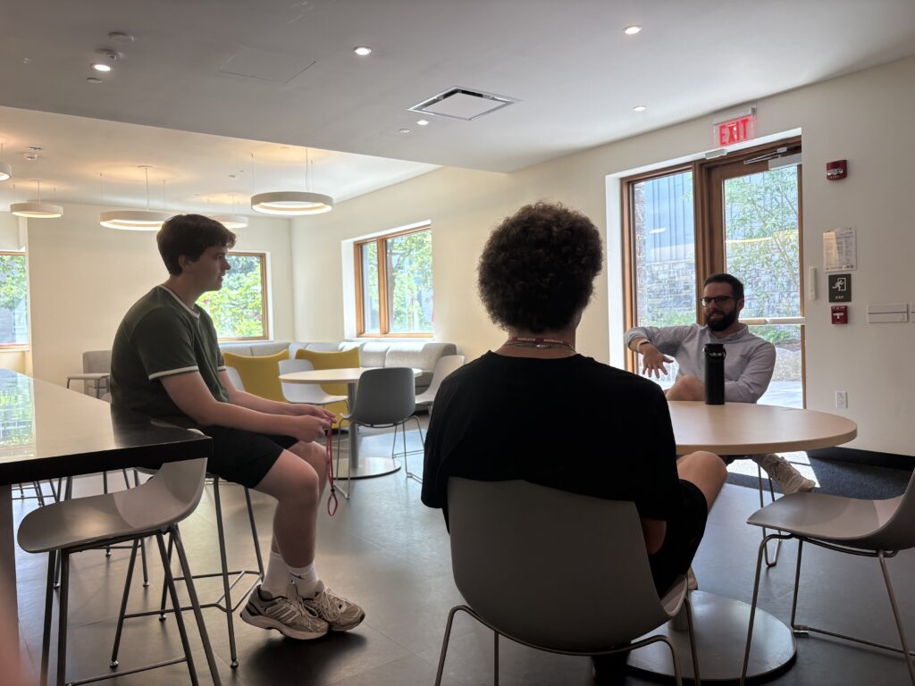 Two students talking at a table with one of the staff professors