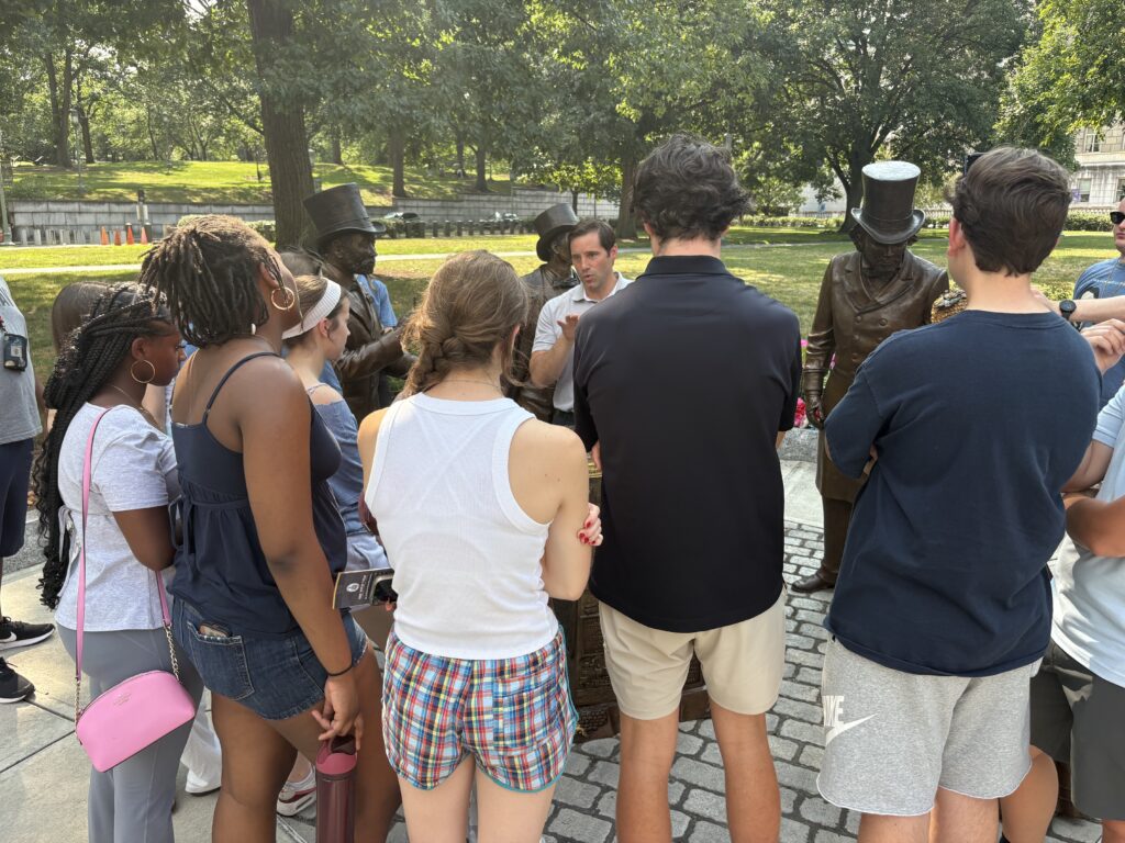 Students gathered around four metal statues, listening to their professor speak in the middle of their formed semicircle 