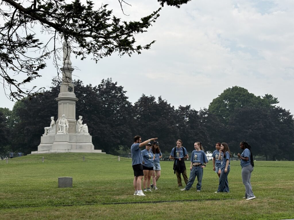 Students standing in front of a large monument, Professor Wingert descriptively talking to them