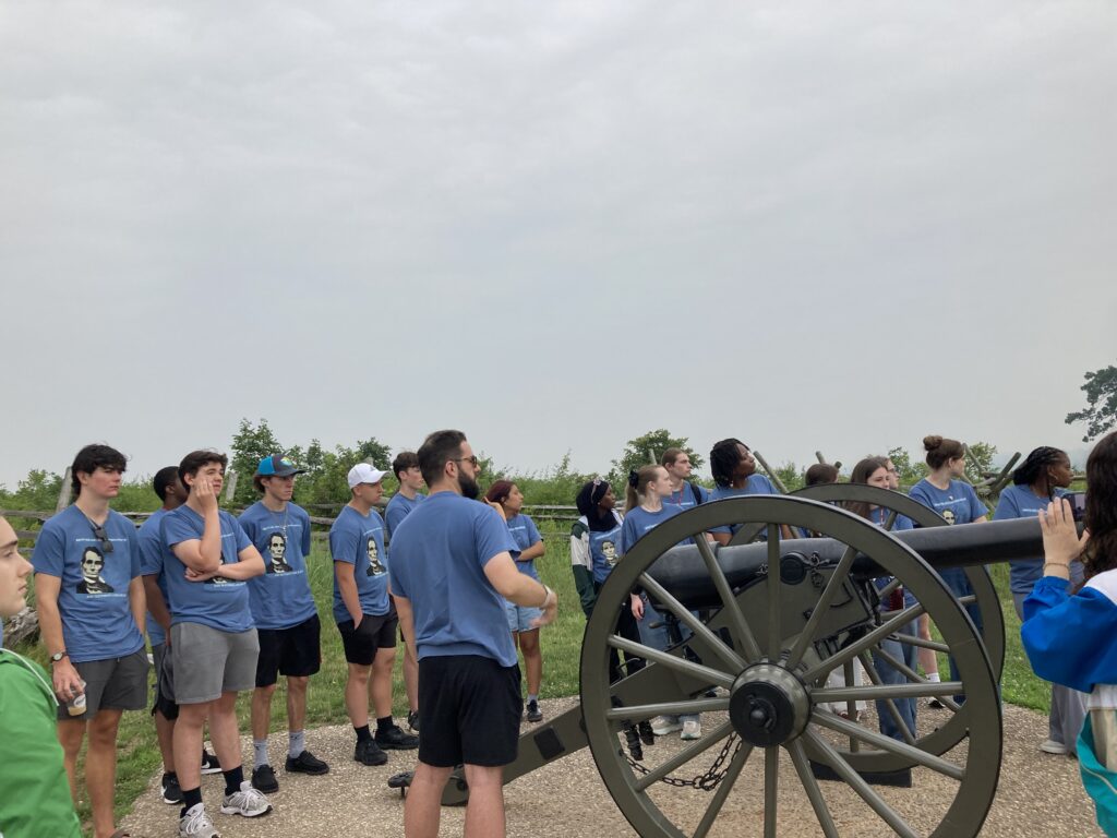 Kids standing in a semi circle around a cannon, listening to Professor Wingert speak, all looking off in the distance