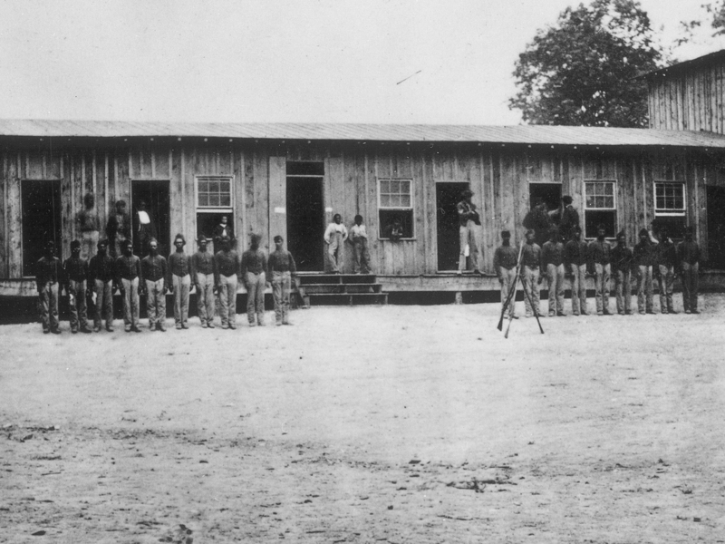 Military barracks constructed out of wood, with uniformed men standing in front. 