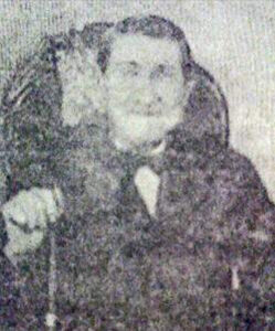 old man seated, holding cane, wooden chair in background